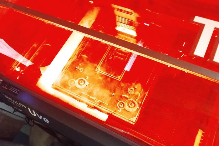 Blick in einen Lasergravierer während der Arbeit: rotes Licht, darunter eine Platte, in die gerade ein Muster gebrannt wird