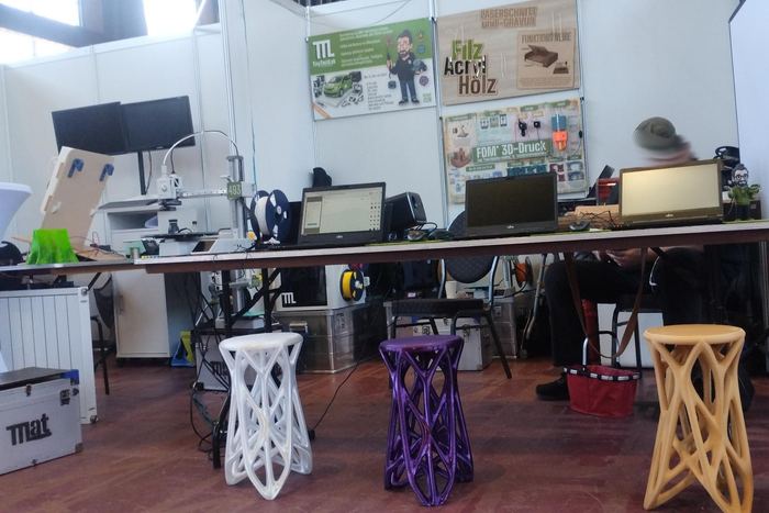 Drei gedruckte Hocker in Weiß, Violett und Holzfarbe vor einem Messestand mit Druckern und Laptops